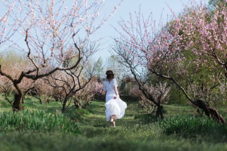 Foto de Young girl in white dress running in a blooming spring peach tree garden - Imagen libre de derechos