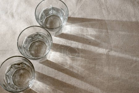 Téléchargez les photos : Crystal glasses with water and sparkling sunlight shadows on neutral beige background. Minimalist aesthetic interior modern design concept - en image libre de droit