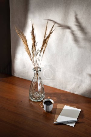 Téléchargez les photos : Esthétique intérieur d'été nature morte avec flic de café, cartes en papier et herbe sèche dans un vase en verre sur une table en bois, ombre de soleil floral sur un mur, concept d'affaires du matin - en image libre de droit