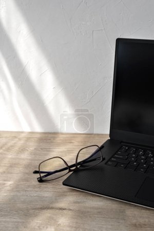 Photo for Minimalist business branding concept, laptop and glasses on a wooden table, geometric sunlight shadows on white wall background, copy space - Royalty Free Image