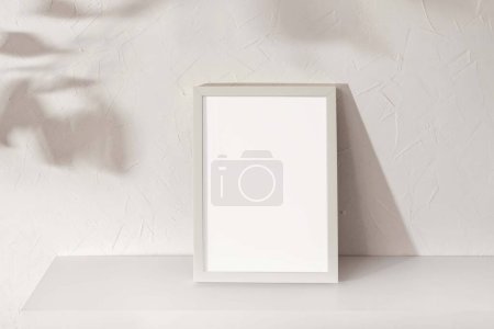Foto de Marco de imagen en blanco con espacio de copia maqueta de pie en estante blanco, sombras de luz solar floral sobre un fondo de pared de hormigón blanco, marca sostenible minimalista o plantilla de diseño de interiores - Imagen libre de derechos