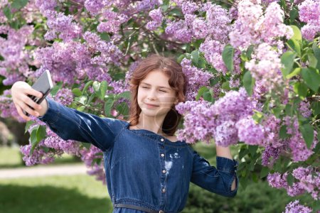 Téléchargez les photos : Jolie adolescente en robe de denim prenant selfie dans un parc fleuri de printemps parmi les fleurs lilas, les yeux fermés - en image libre de droit