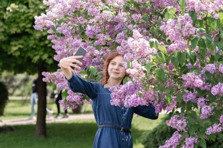 Téléchargez les photos : Jeune adolescente heureuse en robe de denim regardant l'écran du téléphone mobile et prenant un selfie dans le jardin de fleurs lilas, fille prenant photo autoportrait en fleurs pour les médias sociaux ou blog - en image libre de droit