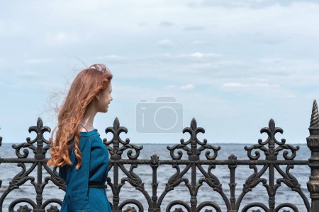 Photo for Cure serene teenager girl with red hair looking at sea and sky nature landscape, enjoying sunny weather and wind breeze outdoor on the embankment - Royalty Free Image