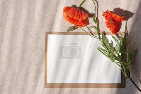 Foto de Hermosa tarjeta de felicitación floral, plantilla de postal con espacio para copiar, tarjeta de papel vacía, sobre, flores de amapola roja sobre un fondo de lino beige neutro con sombras de luz solar - Imagen libre de derechos