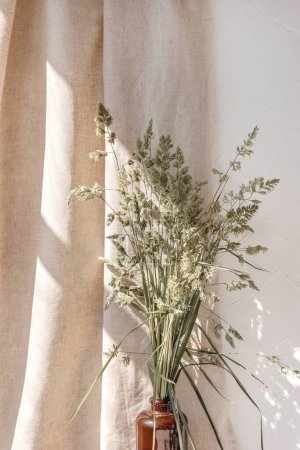 Foto de Estético arreglo interior floral minimalista, hierba verde del prado en jarrón marrón, rayos de sol y sombras sobre un fondo de cortina de lino beige, decoración casera nórdica - Imagen libre de derechos