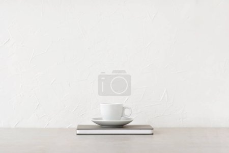 Foto de Taza blanca con bebida y cuaderno sobre una mesa beige neutra, fondo de pared de hormigón blanco con espacio de copia vacío. Plantilla de marca minimalista, coffee break, concepto de almuerzo de negocios, espacio de trabajo de oficina en casa - Imagen libre de derechos