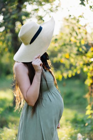 Foto de Mujer embarazada irreconocible en un sombrero de paja y vestido verde, caminando en la naturaleza al aire libre, concepto de embarazo, salud de la maternidad, estilo de vida eco sostenible, fertilidad y crianza consciente. - Imagen libre de derechos
