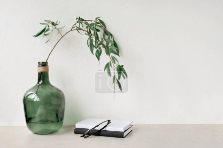 Foto de Moderna botella de vidrio verde con rama de sauce, libros y vasos sobre una mesa beige. Fondo de pared de hormigón beige neutro vacío con espacio para copiar. Plantilla interior escandinava minimalista - Imagen libre de derechos