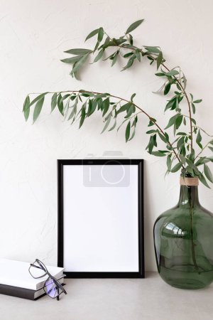 Photo for Empty black picture frame mockup, green glass vase with tree branch, stationery on a beige tabletop and wall background. Minimalist sustainable neutral interior decoration, art poster template - Royalty Free Image