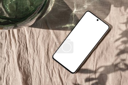 Foto de Maqueta de pantalla de teléfono inteligente vacía en un mantel de lino beige con sombra de luz solar estética, plantilla de marca de negocio de estilo de vida - Imagen libre de derechos