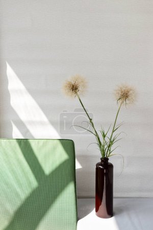 Foto de Ramo de flores de diente de león en jarrón en la mesa, silla verde, sombra de luz solar estilo de vida sobre un fondo de pared vacío neutro. Interior rústico simple mínimo, estilo de vida sostenible, concepto de vida lenta - Imagen libre de derechos