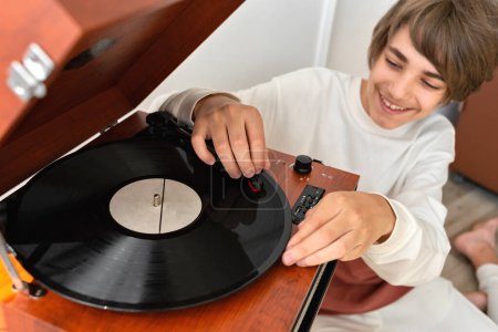 Photo for Happy smiling handsome teenager boy turning on retro vinyl player, brown wooden turntable and listening music - Royalty Free Image