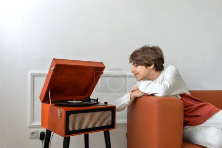 Foto de Guapo adolescente escuchando discos de vinilo en un tocadiscos de madera retro, acostado en un sofá marrón y sonriendo, disfruta del sonido de la música - Imagen libre de derechos