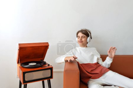 Photo for Happy beautiful teenager boy sitting on a brown sofa, listening vinyl record on retro wooden turntable, smiling, enjoying music, having fun. Empty white wall background - Royalty Free Image