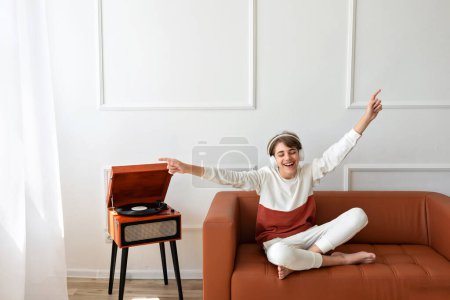 Photo for Happy beautiful teenager boy sitting on brown sofa with hands rising and eyes closed, listening vinyl record on retro wooden turntable, smiling,enjoying music, having fun, empty white wall background - Royalty Free Image