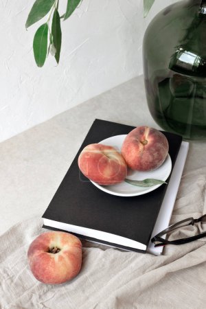 Foto de Bodegón estético de verano, fruta madura del melocotón, libro, vasos, jarrón verde con rama de árbol sobre un mantel de lino, fondo blanco vacío de la pared - Imagen libre de derechos