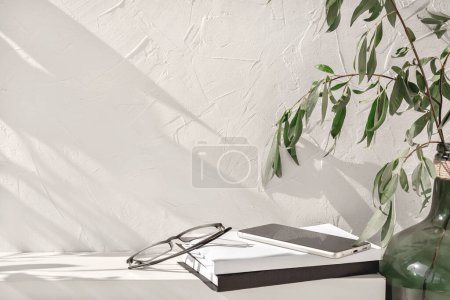 Photo for White bookshelf with books, glasses, mobile phone, green vase with plants. Empty concrete wall background with aesthetic floral sunlight shadows. Shelf, podium stage mockup, home interior template - Royalty Free Image