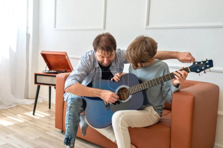 Foto de Padre soltero enseñando hijo adolescente a tocar una guitarra acústica en interiores - Imagen libre de derechos