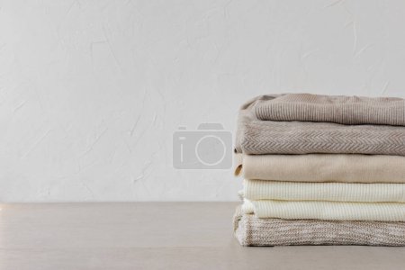 Photo for Wool knitted clothes stack closeup in a neutral beige colors on table on white empty wall background. Aesthetic fashion minimalist wardrobe concept, warm autumn knitwear jersey - Royalty Free Image