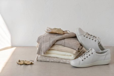 Photo for Fashion minimalist autumn clothes wardrobe in neutral beige colors. Beige, creamy, taupe wool knitted sweaters stack, white sneakers, brown fall leaves on table, on a white empty wall background - Royalty Free Image