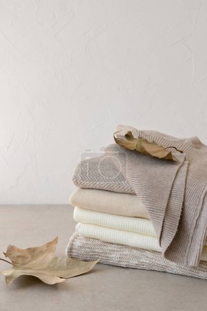 Photo for Early autumn fashion clothes concept. Minimalist neutral beige knitted sweaters stack, fall leaves, wardrobe for september. Empty white wall background, copy space. - Royalty Free Image