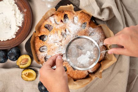 Téléchargez les photos : Décoration prune torte avec glaçage. Mains tenant tamis avec poudre de sucre au-dessus de tarte aux prunes, ce qui rend boulangerie tarte aux fruits. - en image libre de droit