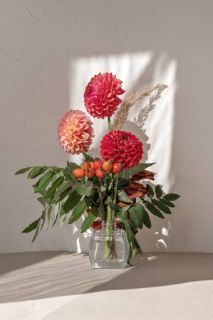 Téléchargez les photos : Bouquet de fleurs d'automne festif avec dahlia rouge et rose et baies orange dans un vase sur la table avec ombre solaire esthétique - en image libre de droit