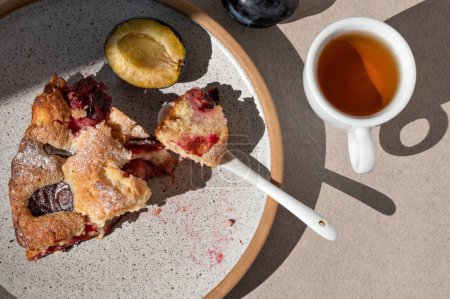 Photo for Piece of plum torte on a plate, cup with tea, teaspoon on neutral beige table with sunlight shadow. Lifestyle morning breakfast, sliced fruit pie, cake - Royalty Free Image