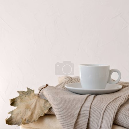 Téléchargez les photos : Tasse blanche de boisson au café chaud, pull tricoté taupe beige pastel, feuille d'automne séchée sur un fond neutre et vide. Modèle minimaliste de conception de marque ou de blog d'automne esthétique, espace de copie - en image libre de droit