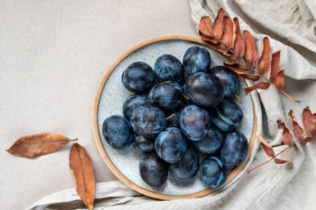 Foto de Frutas maduras de ciruela en plato sobre mesa beige neutro y fondo de mantel de lino con hojas secas de otoño. - Imagen libre de derechos