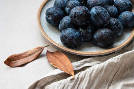 Téléchargez les photos : Fruits de prune sur table avec gouttes d'eau, nappe en lin et feuilles d'automne brunes sur fond de table beige neutre. - en image libre de droit