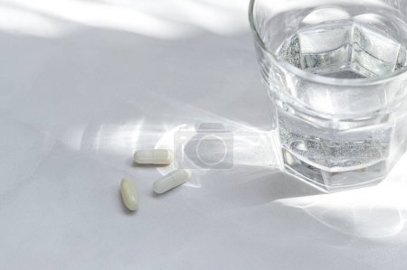 Téléchargez les photos : Pilules et capsules blanches, verre avec de l'eau pure sur fond de table blanche avec des ombres de lumière naturelle du soleil. Concept minimal de soins de santé et de médecine esthétique, gros plan. - en image libre de droit