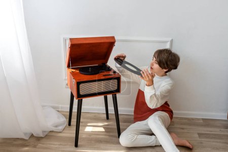 Photo for Young boy preparing listening music. Handsome teenager sitting on floor, putting the vinyl record in retro wooden brown turntable, player. Minimalist white home interior. - Royalty Free Image