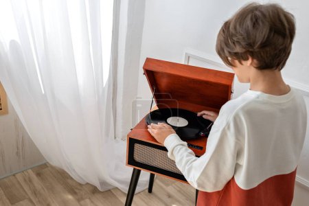 Téléchargez les photos : Beau garçon adolescent mettant disque vinyle dans une platine en bois brun rétro, lecteur, préparation de musique d'écoute. Vue arrière, intérieur minimaliste - en image libre de droit