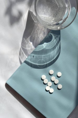 Téléchargez les photos : Pilules blanches, carnet bleu clair, verre avec de l'eau sur la table blanche avec des ombres de soleil esthétiques. Concept de santé et de médecine. - en image libre de droit
