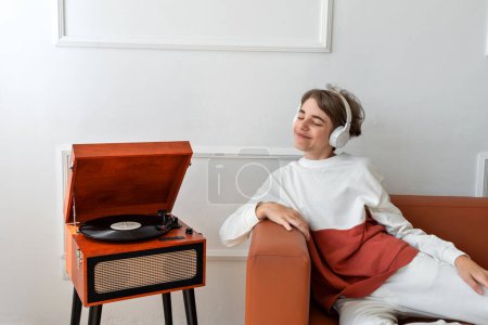 Photo for Beautiful teenager boy wearing earphones smiling and listening music near wooden vinyl record player. Minimalist home interior, retro aesthetic. - Royalty Free Image