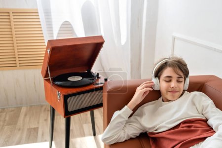 Photo for Teenager boy with eyes closed chilling, laying on sofa and listening music in earphones. Wooden turntable, vinyl record player standing behind. - Royalty Free Image
