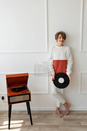 Photo for Retro vinyl record music trend aesthetic. Handsome teenager boy standing near wooden brown turntable, holding vinyl record in hands and smiling. Home interior, white wall background. - Royalty Free Image