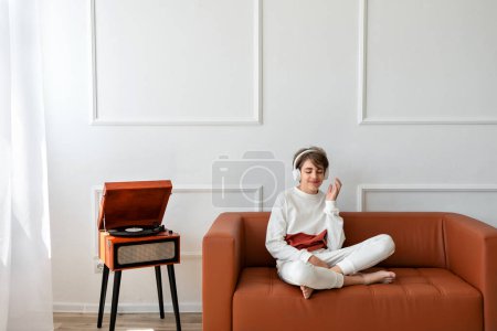 Foto de Feliz adolescente sonriente sentado en un sofá con auriculares y escuchando música cerca del tocadiscos con disco de vinilo. - Imagen libre de derechos