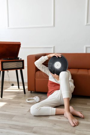 Photo for Teenage boy sitting on floor with vinyl record in hands, hiding face behind. Turntable on background. - Royalty Free Image