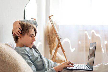 Foto de Adolescente serio mirando la pantalla del portátil, tecleando en el teclado, sentado en la cama. Estudio en línea, concepto de educación a distancia. - Imagen libre de derechos