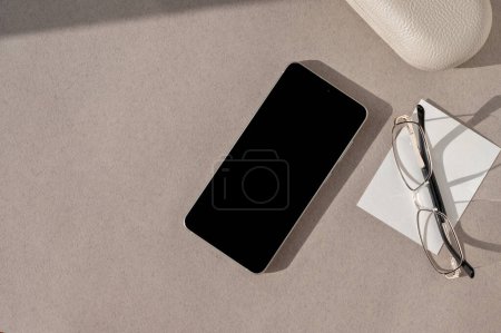 Photo for Minimal business brand template, flat lay. Mobile phone with empty black screen, eyeglasses, paper card on neutral beige background with natural sun light shadow. - Royalty Free Image