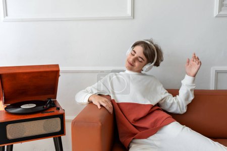 Foto de Lindo adolescente con auriculares escuchando música y divirtiéndose, relajándose en un sofá cerca de un tocadiscos de madera con grabación de vinilo. - Imagen libre de derechos