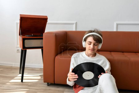 Photo for Teenager boy with headphones holding in hands vinyl record and looking on it. Brown wooden turntable and sofa on background, home interior, lifestyle. - Royalty Free Image