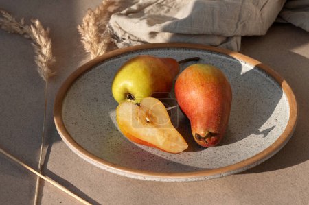 Téléchargez les photos : Ripe whole and sliced half red pears on plate on beige table background in natural sunlight with shadows. Aesthetic lifestyle autumn food still life. - en image libre de droit