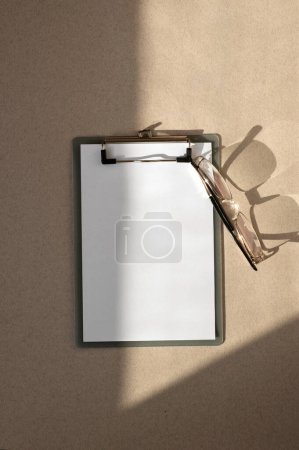 Téléchargez les photos : Modèle de marque d'entreprise minime, maquette de feuille de papier vierge sur presse-papiers et lunettes sur fond de bureau beige avec ombre solaire naturelle. - en image libre de droit