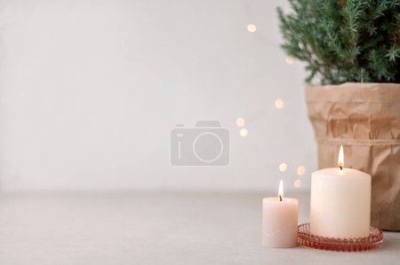 Téléchargez les photos : Bougies et guirlandes floues déconcentrées, genévrier en pot sur table beige et fond blanc vide. Décoration de maison de Noël scandinave esthétique, modèle de design d'intérieur. - en image libre de droit