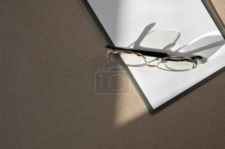 Photo for Minimal business brand template. Clipboard with white paper sheet closeup, eyeglasses on brown table with natural sunlight shadows background. - Royalty Free Image