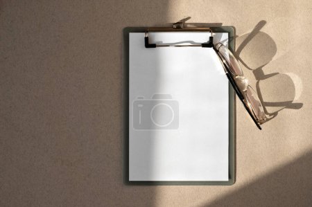 Photo for Clipboard with blank white paper sheet mockup, eyeglasses, aesthetic lifestyle sunlight shadows on neutral beige desk background. Elegant business branding template, flat lay. - Royalty Free Image